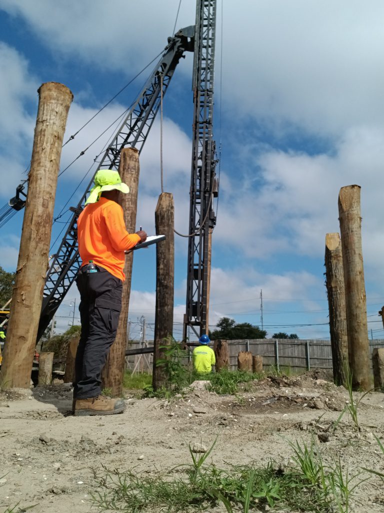 Construction workers operating machinery on a building site with large wooden posts.